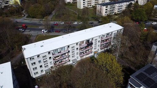 Luftaufnahme eines langgestreckten, mehrgeschossigen Gebäudes mit weißem Flachdach und Balkonen