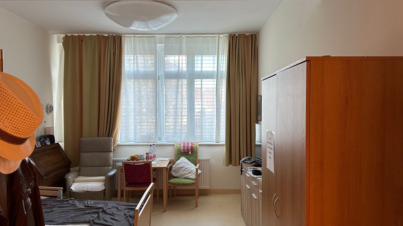 Zimmer mit Bett, Sessel, Tisch und Holzschrank; Fenster mit weißen Gardinen und seitlichen Vorhängen