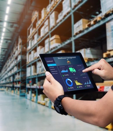 Hände bedienen ein Tablet mit einem Warehouse-Management-Dashboard vor Hochregalen in einer Lagerhalle